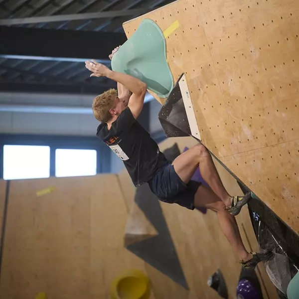 Een man met kort blond haar beklimt een indoor-bouldermuur; hij houdt zich vast aan een grote turquoise greep terwijl hij met gebogen benen hangt en met zijn voeten tegen de muur duwt. Hij draagt een zwart shirt, een marineblauwe korte broek en klimschoen
